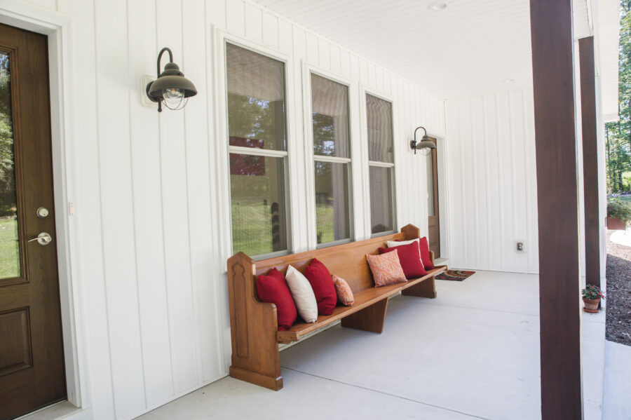 8 foot deep front porch with stained wood square posts and reddish-pink pillows on a wooden bench. 