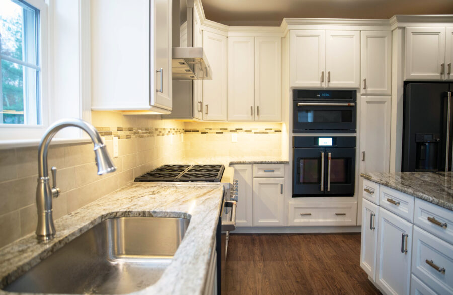 The kitchen's large single bowl stainless steel sink with tiled backsplash, stove, and oven with white cabinets. 