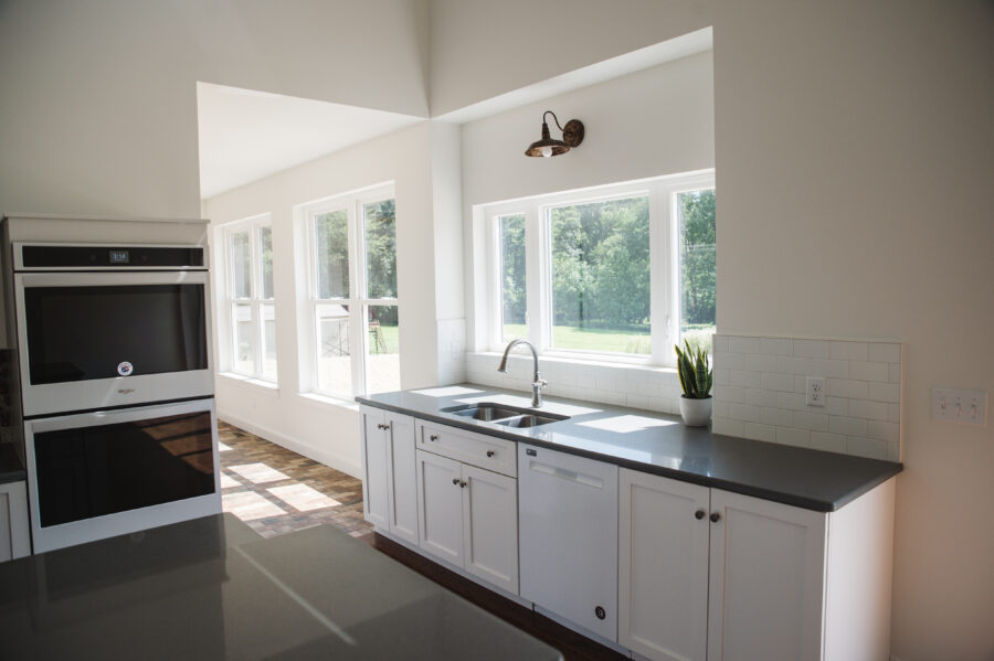 The large window in the kitchen behind the kitchen sink on the rear wall with white cabinets and walls and a quartz countertop 